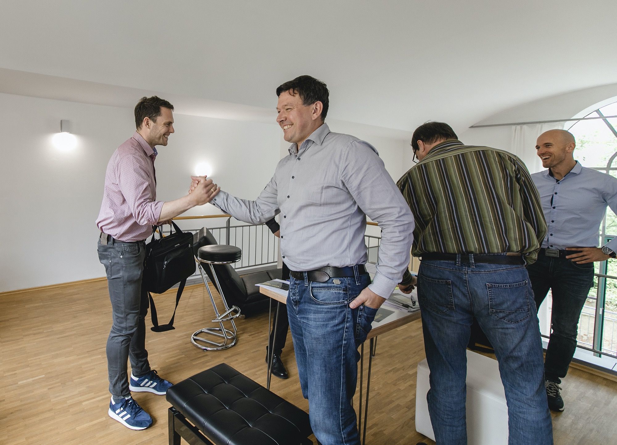 Zwei Teilnehmer begrüßen sich per Handschlag und lachen, im Hintergrund weitere Teilnehmer im Gespräch bei einem Workshop